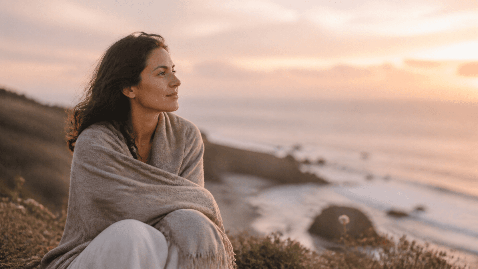 A woman wrapped in a soft blanket looking peacefully across the ocean at sunset